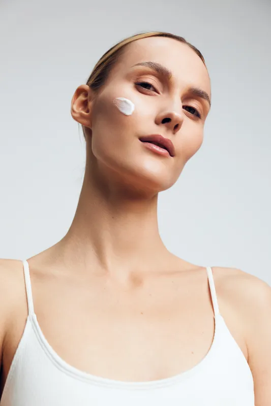 A young woman with a dollop of cream on her cheek looks at the camera in a minimalist studio. Close-up portrait focusing on skincare, beauty routine, and self care concepts.