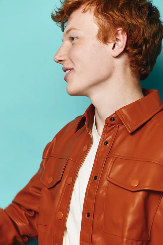 Portrait of young redhead man wearing leather jacket with white shirt, smiling looking sideways on blue background. Casual style with natural expression and profile view.