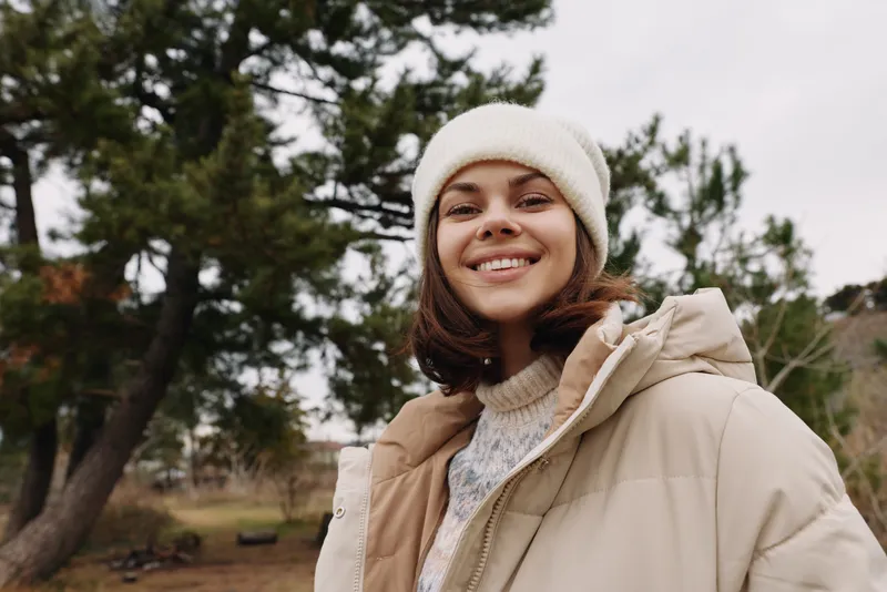 Woman outdoors in a warm beige coat and knit beanie smiles at the camera in a park with trees and open space, showcasing autumn calm and casual style.
