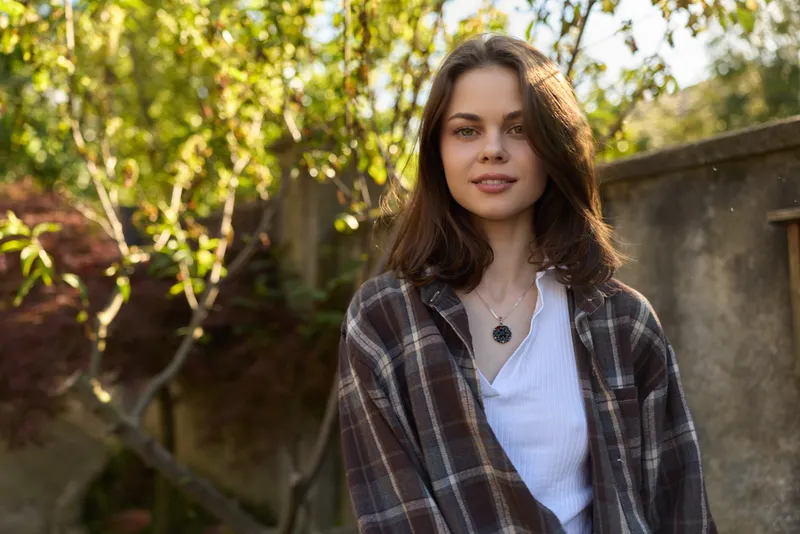 A casual outdoor portrait of a young woman in a plaid coat, smiling softly in warm daylight. The scene conveys relaxed lifestyle and natural confidence, suitable for everyday fashion and outdoor
