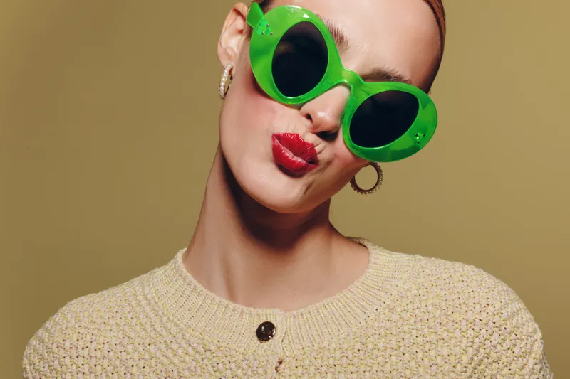 Young woman with bright red lipstick wearing oversized green sunglasses and beige sweater poses with puckered lips on neutral background. Concept of fashion, style, and playful attitude.