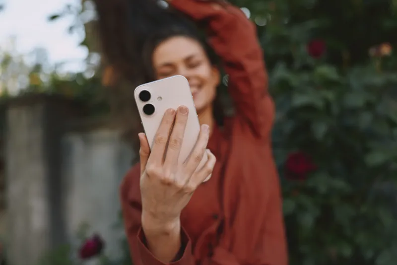 Cheerful woman outdoors, taking a selfie with a smartphone, warm sunlight, relaxed pose, casual outfit, natural colors, leafy background, candid lifestyle moment