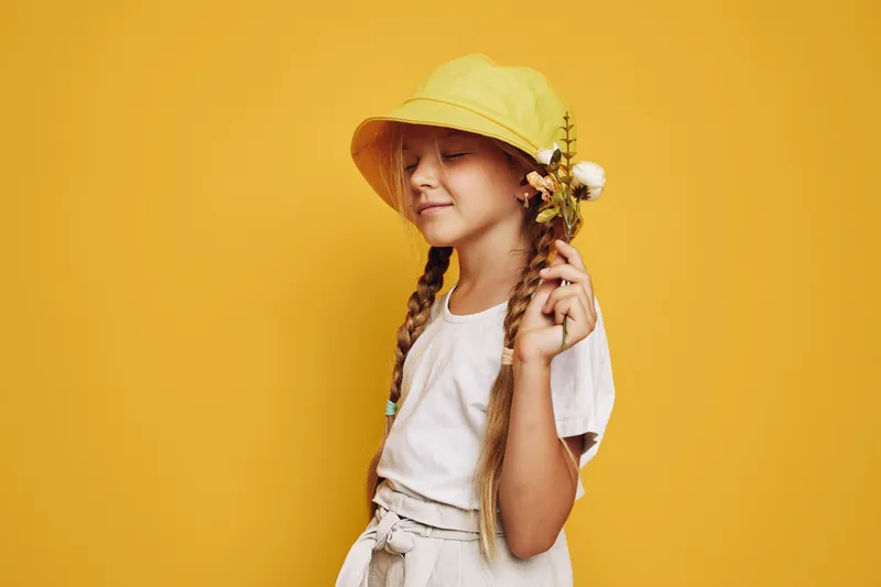 Young girl with braided hair wearing a yellow hat holds wildflowers against a yellow background. She looks peaceful and serene with closed eyes.