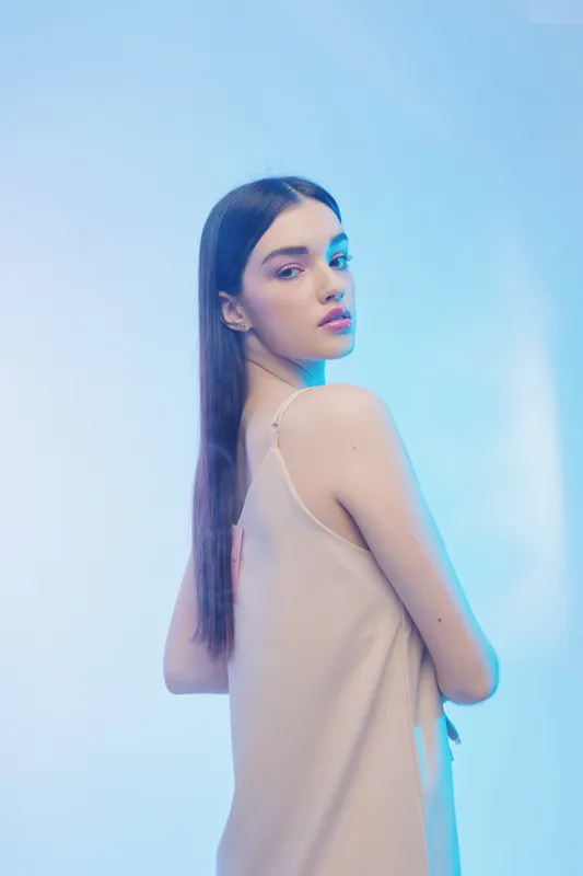Young woman with long dark hair poses in a beige dress against a light blue background. Her makeup highlights her natural beauty and fashion style. Studio portrait.