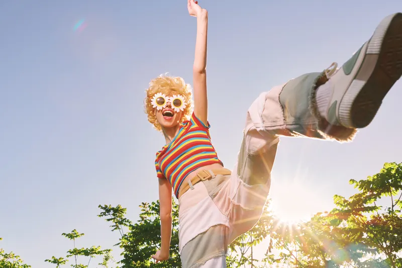 Young woman with curly hair leaps outdoors in a striped shirt and sunglasses, smiling broadly as sunlight bursts through trees in a vibrant retro moment with warm tones.