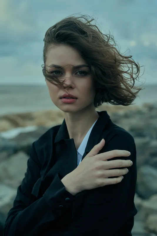 Woman stands by the sea in windy weather, wearing a dark blazer, with a thoughtful gaze and tousled hair, capturing moody coastal fashion and emotional depth.