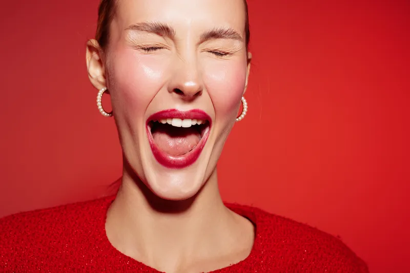 Close-up of a happy woman with red lipstick and earrings laughing with closed eyes on red background. Concept of joy, beauty, emotion, and vibrant fashion portrait.