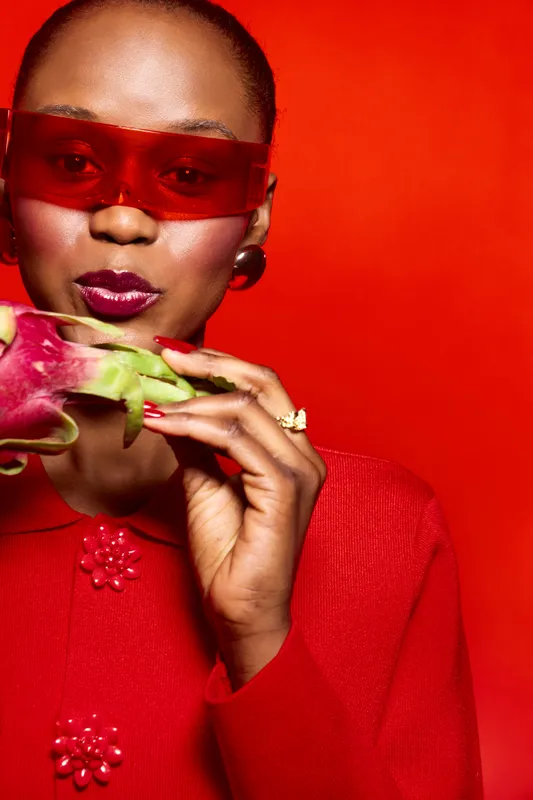 Stylish dark skinned African woman in red sweater and red sunglasses holds a dragon fruit. Bright red background enhances vibrant fashion and fresh fruit concept in studio portrait.
