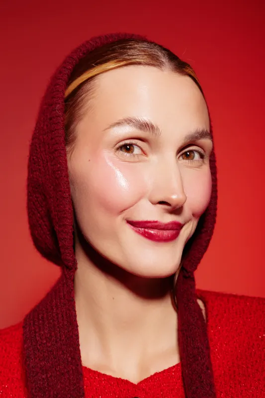 Close-up portrait of a smiling woman with glossy skin wearing a red knitted hood and sweater against a red background. Concept of beauty, fashion, winter style, and confident expression.