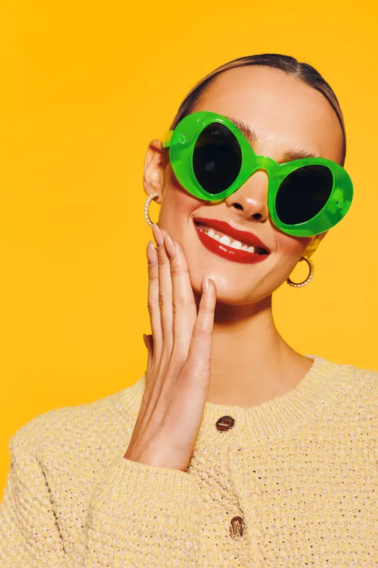 Smiling woman with bright red lipstick wearing large green sunglasses and yellow textured sweater poses against vibrant yellow background. Concept of fashion, summer style, and confidence.