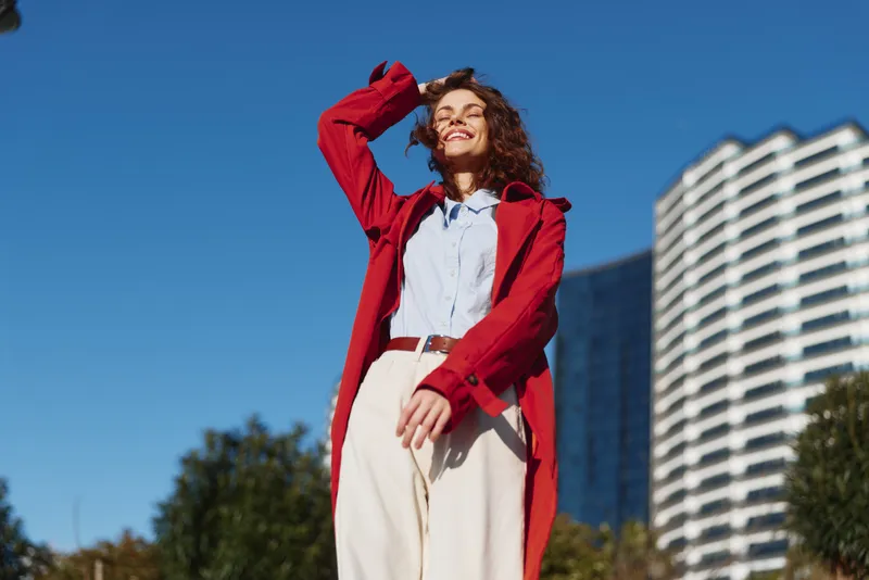 Young woman with curly hair enjoys a sunny day outdoors wearing a red coat and white pants with a joyful expression against a clear blue sky and urban background.