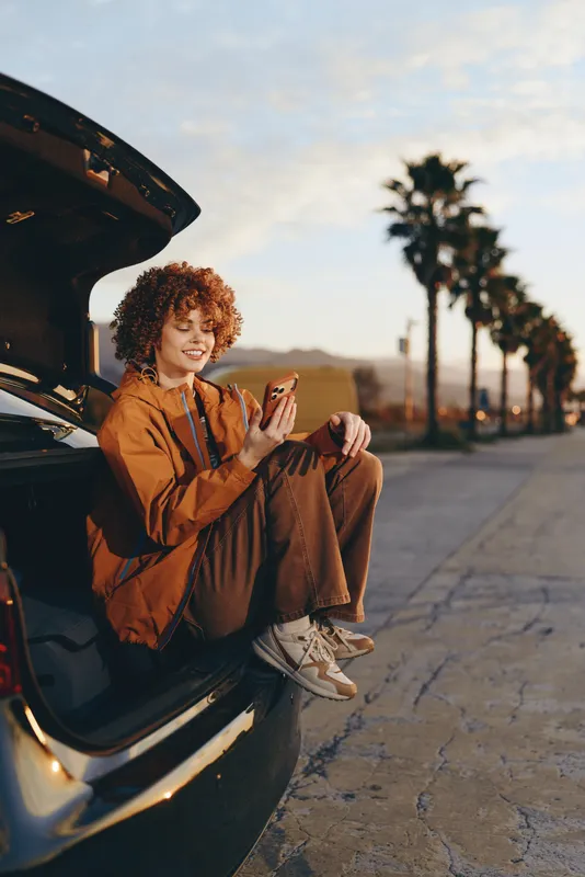 Cheerful woman lifestyle outdoors, smiling by open car trunk on a sunlit roadside, curly hair, casual jacket and jeans, sunset sky and palm trees in the background, soft warm light