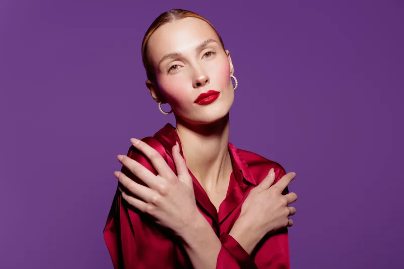 Portrait of a confident woman with slicked back hair wearing red satin blouse and bold red lipstick. She poses with crossed arms against a vibrant purple background. Fashion and beauty concept.