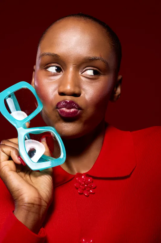 Close-up portrait of a dark skinned African woman with short hair holding turquoise eyeglasses near her face, wearing a red top, looking to the side with a playful expression on red background.