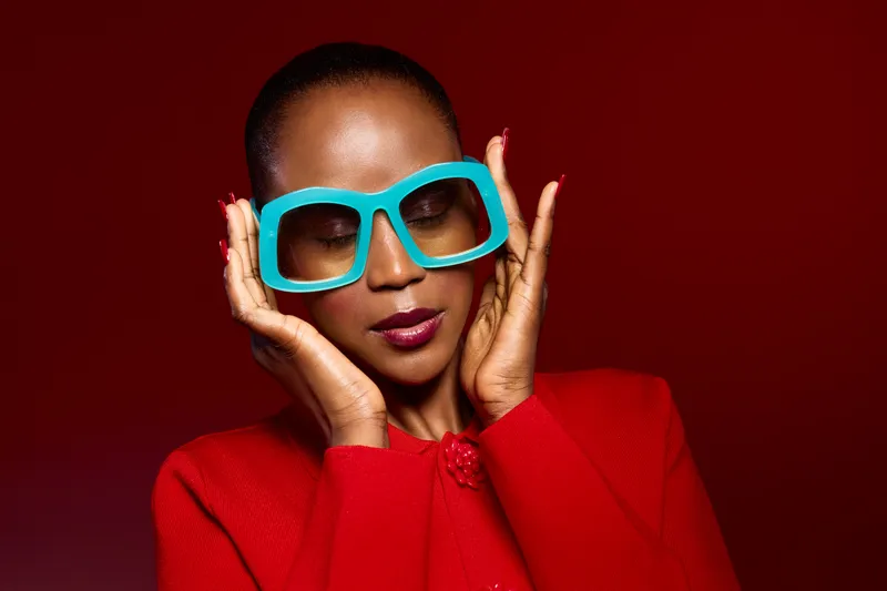 Portrait of a confident African woman with dark skin wearing oversized blue glasses and a red outfit. Studio shot with red background, highlighting fashion and style concepts.