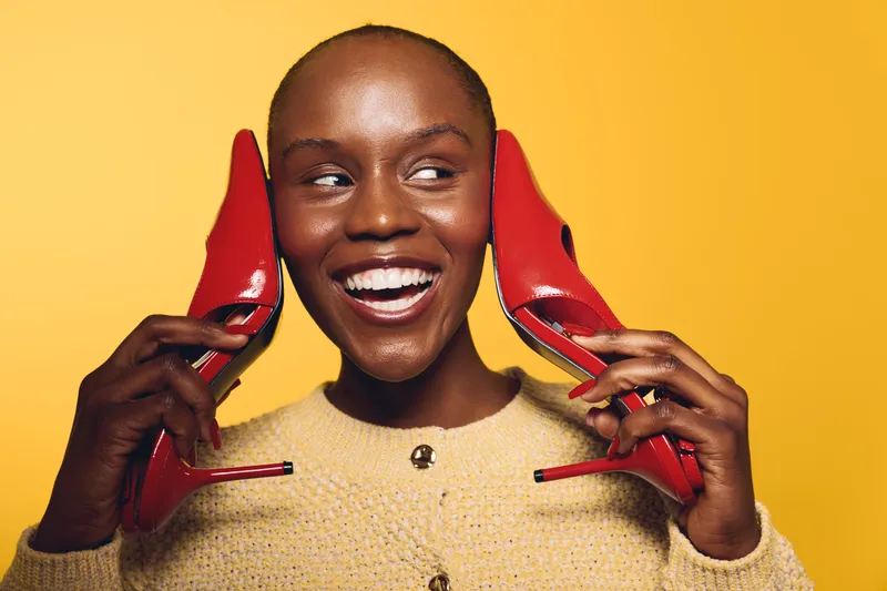 Smiling black woman with dark skin holds red high heels near face on yellow background. Fashion concept with joyful African female model wearing beige sweater and bright accessories.