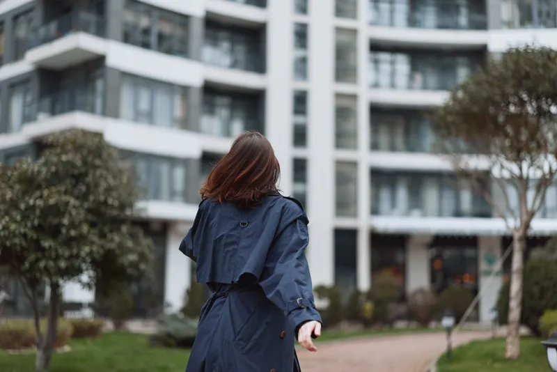 A woman with shoulder length brunette hair walks away in a navy trench coat through a modern apartment complex courtyard.