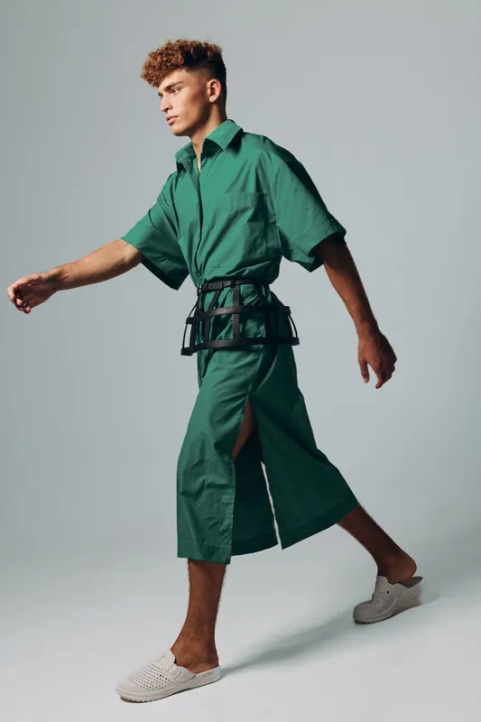 Young man walking in green oversized shirt and matching wide pants with side slit, wearing white slip-on shoes and black belt accessory on plain background in studio.