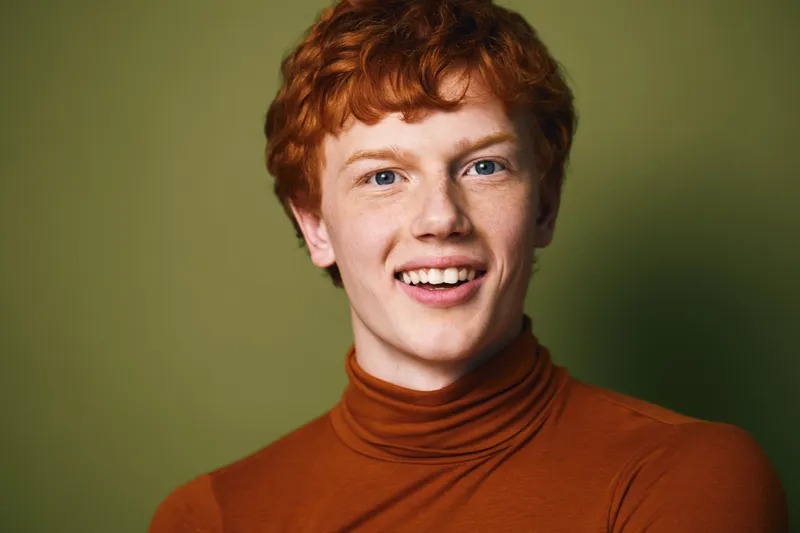Young man with red hair smiling and wearing brown turtleneck sweater posing against green background with confident and happy expression in studio portrait.