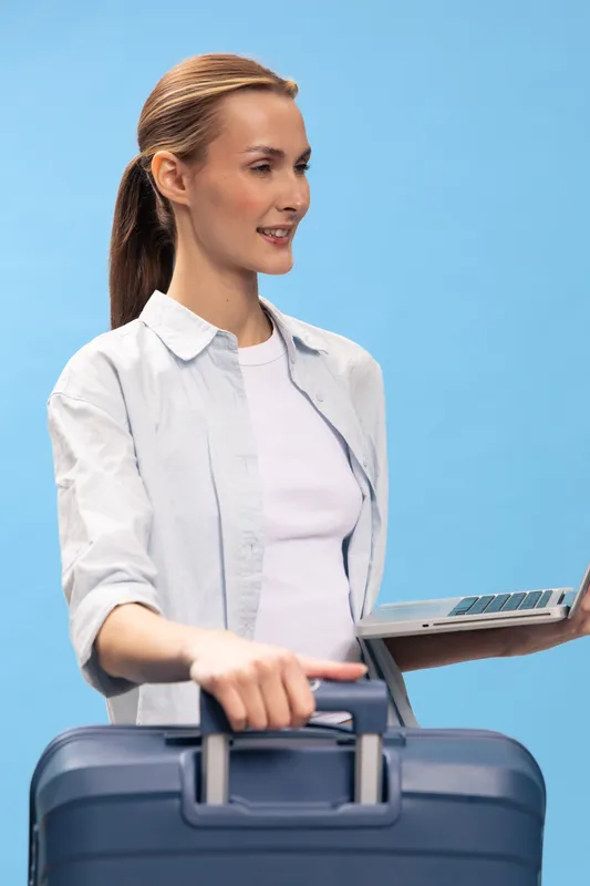 Young woman traveler with suitcase and laptop stands against blue background, smiling and ready for business trip or vacation. Concept of travel, technology, and planning.