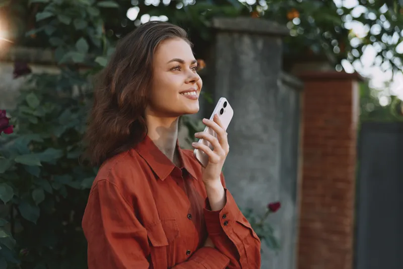 Young woman wearing a rust red shirt smiles softly while talking on a smartphone in a leafy garden, capturing a casual lifestyle moment outdoors and relaxed