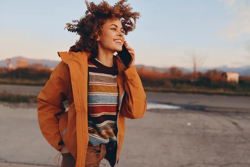 Woman lifestyle portrait, outdoors at autumn sunset, curly haired woman in orange jacket and striped sweater, smiling while walking on rural road, casual travel atmosphere, warm evening light