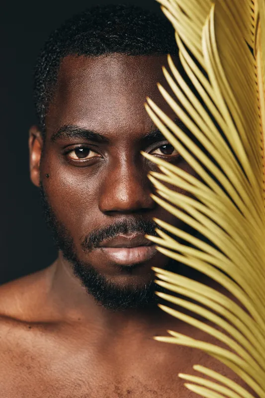 African man with dark skin and short hair posing with palm leaf in creative conceptual visual style. Closeup portrait with serious expression and natural lighting on face and shoulder.