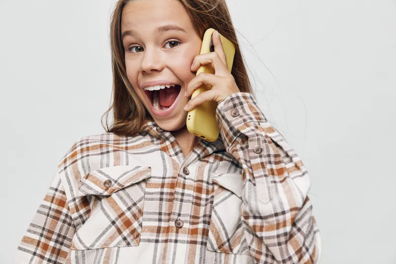 Girl in plaid shirt smiling with phone near ear talking happily against white background in bright studio. Youth, communication and emotions with cheerful expression and casual style.