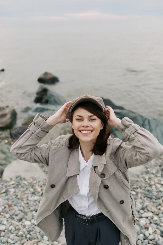 Smiling young woman in patterned coat and cap stands on rocky beach by sea, holding hat with both hands. Casual fashion and outdoor lifestyle concept with natural light and relaxed mood.