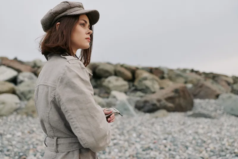 Young woman in beige coat and hat stands on rocky beach looking thoughtfully at distance under overcast sky. Concept of contemplation, autumn, and natural outdoor environment.