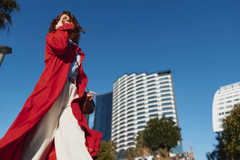 Young woman with curly hair wearing a long red coat walks confidently in an urban setting under clear blue sky during daytime.