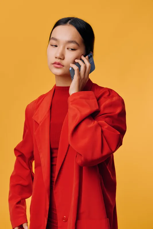 Young woman in red jacket and top holding smartphone near face talking on phone with serious expression on yellow background studio portrait