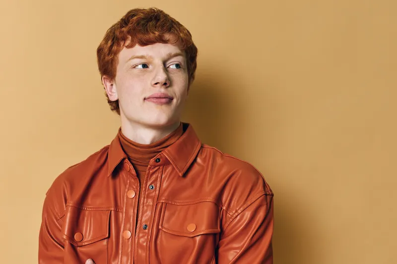 Young man with red hair in a brown leather jacket and turtleneck looks sideways on beige background. Portrait of confident male with casual style and thoughtful expression in studio setting.