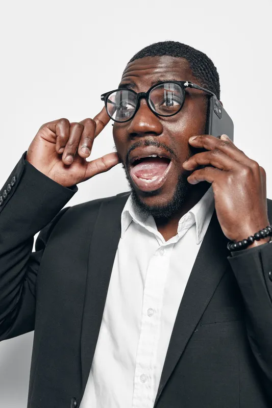 African American man with dark skin wearing glasses and black suit talking on smartphone with surprised expression isolated on white background.