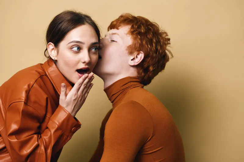 Woman shares a secret whisper to surprised female friend with hand covering mouth on beige background. Two young adults in casual clothing expressing emotion and connection.