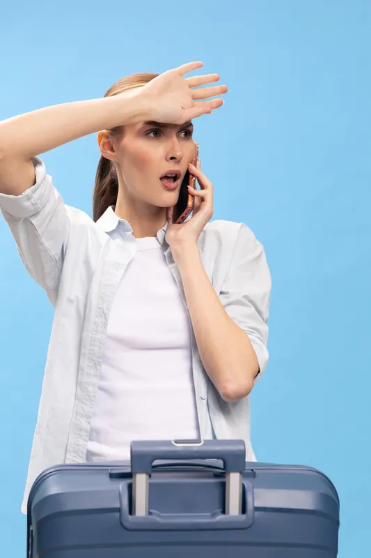 Surprised young woman with suitcase talks on phone, shielding eyes with hand. Travel concept with unexpected situation, trip preparation, and urgent communication on blue background.