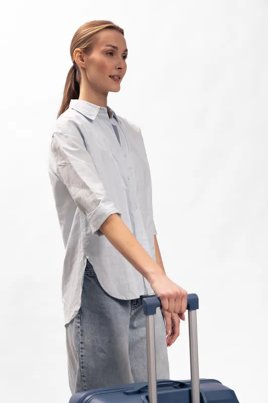 Young woman with long hair and casual shirt holding blue suitcase handle, standing against white background. Concept for travel, tourism, and vacation preparation.