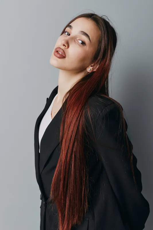 Young woman with long red hair wearing black blazer and white shirt posing confidently against gray background. Portrait of stylish female model with natural makeup and earrings indoors.