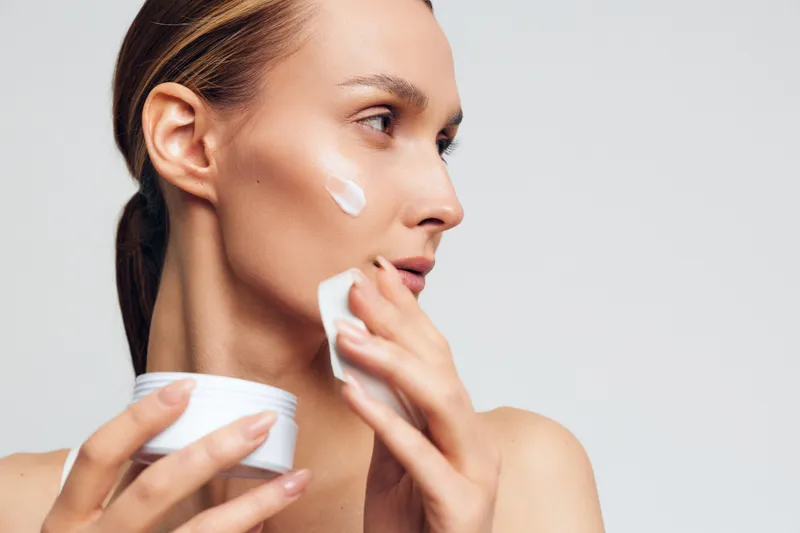 Woman skincare cream face beauty close up moisturizer hygiene spa, applying cream to face with cotton pad, calm studio moment, soft light, smooth skin, white background, care