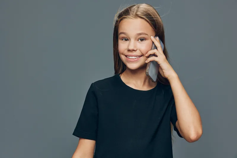 Girl smiling and talking on smartphone with hand near ear. Child in casual black t shirt standing on gray background with happy expression during mobile call.