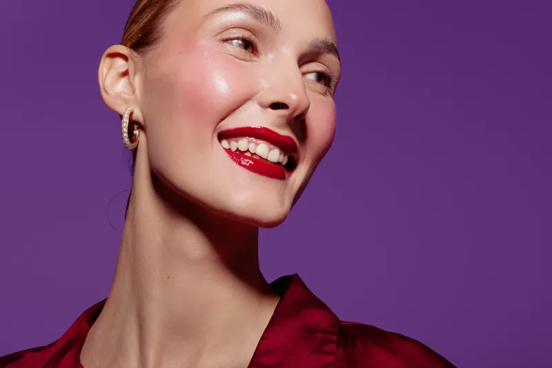 Smiling woman with red lipstick and earrings looks away on purple background. Close-up portrait with smooth skin and shiny makeup. Concept of beauty, fashion, and confidence.