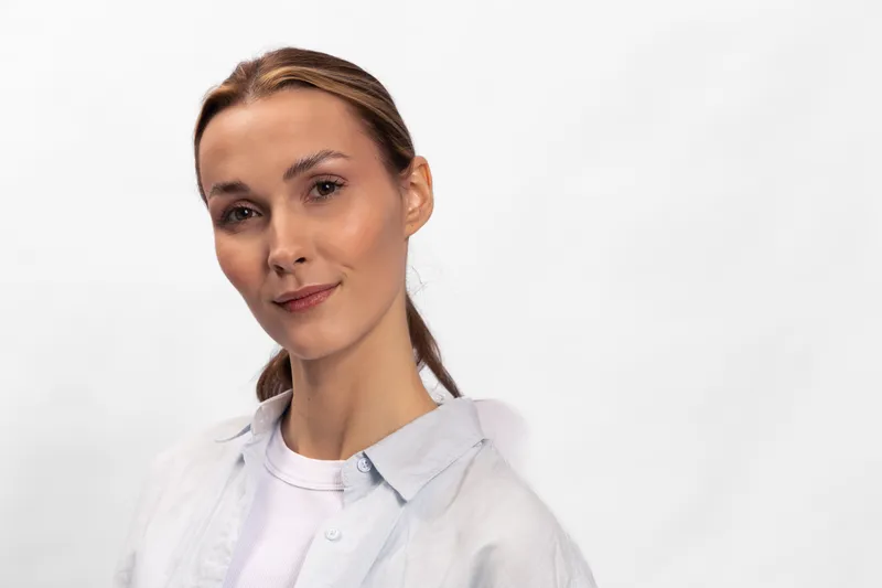 Confident young woman with light makeup and tied hair wears casual shirt over white top, posing against plain white background with slight smile. Concept of natural beauty and casual fashion.
