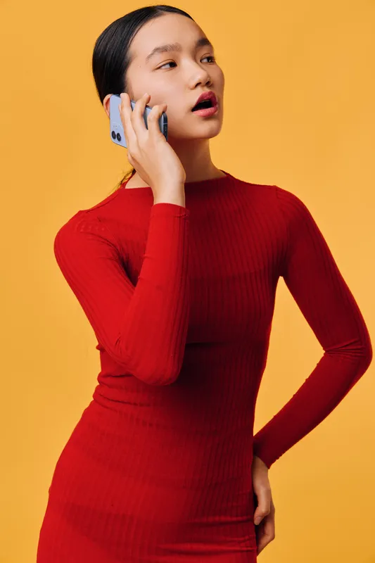 Portrait of young woman with dark hair in red dress talking on smartphone, standing against yellow background. Communication, technology and casual style with confident expression and hand on hip.