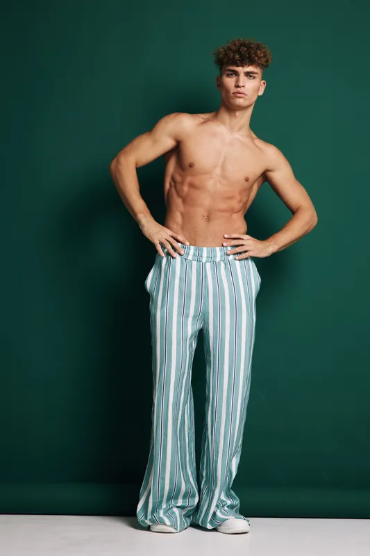 Young man standing shirtless with hands on hips wearing wide stripped pants against green backdrop. Confidence, casual pose and athletic body frame in fashion studio setting.