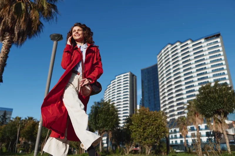 Young woman in a red trench coat walks through a sunny urban park with tall buildings behind, exuding confidence and city vibes as her white pants billow and a crossbody bag sways.