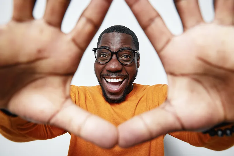 African American man with dark skin and glasses smiling and framing face with hands in orange sweater on white background. Happy, joyful expression and casual style.