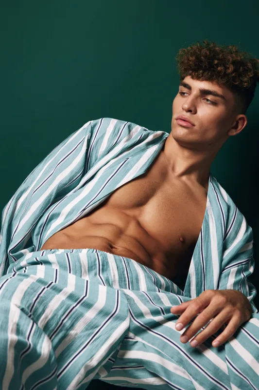 Young man with curly hair wearing striped casual suit without shirt poses reclining against dark green backdrop studio frame confident fashion lifestyle model summer style.