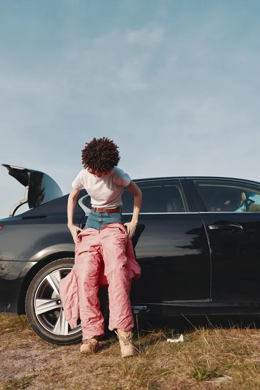 Person adjusting pink pants beside a sleek car in an open field, casual streetwear style under a clear blue sky, relaxed fashion with outdoor ambiance.