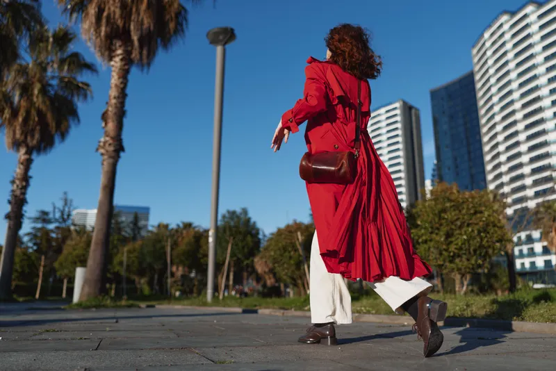 A woman in a red coat walks across a city park with tall buildings in the background, carrying a brown handbag and captured in motion.