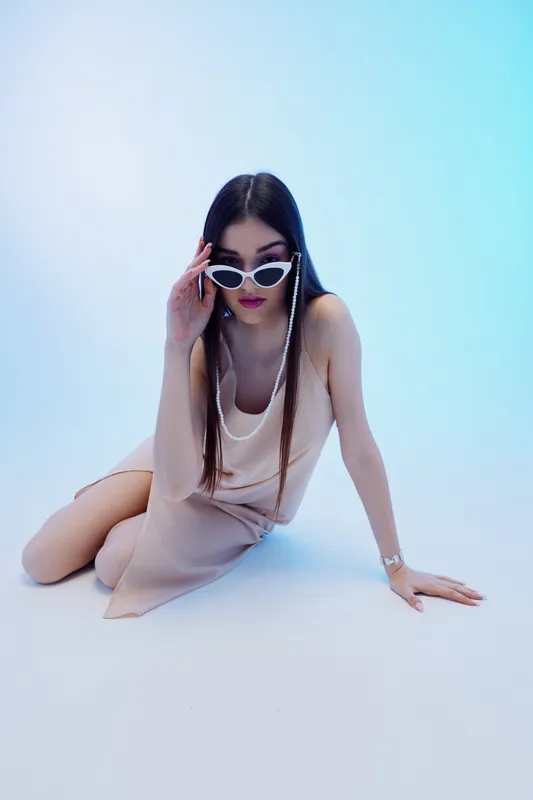 Young woman in fashionable beige dress and white sunglasses poses on floor with blue background. Concept of woman beauty fashion and modern style.